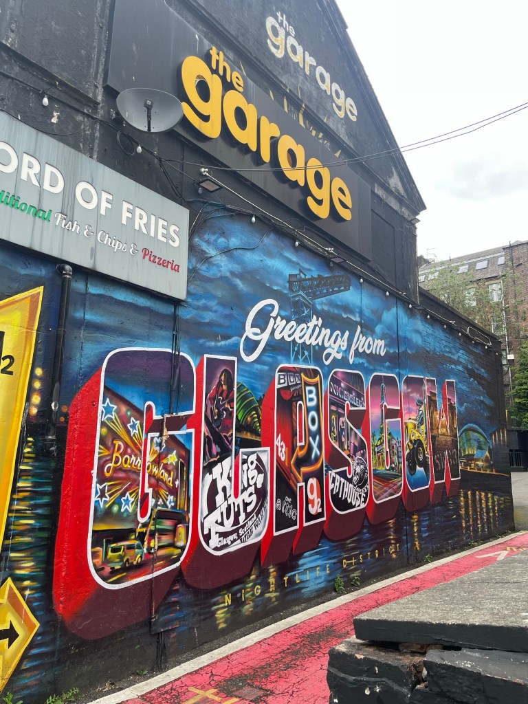 A colorful mural on the side of a building featuring the text 'Greetings from Glasgow,' with artistic representations of local attractions and vibrant imagery.