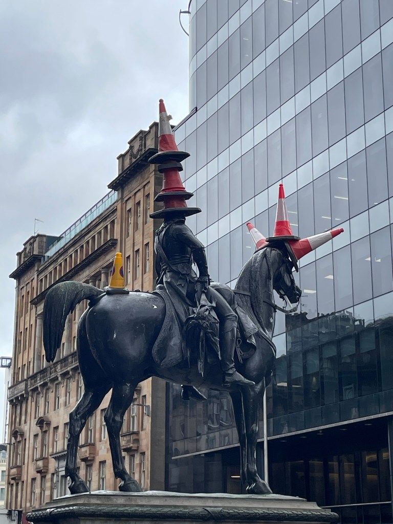 A statue of a mounted soldier wearing multiple traffic cones on its head, with a modern building in the background.