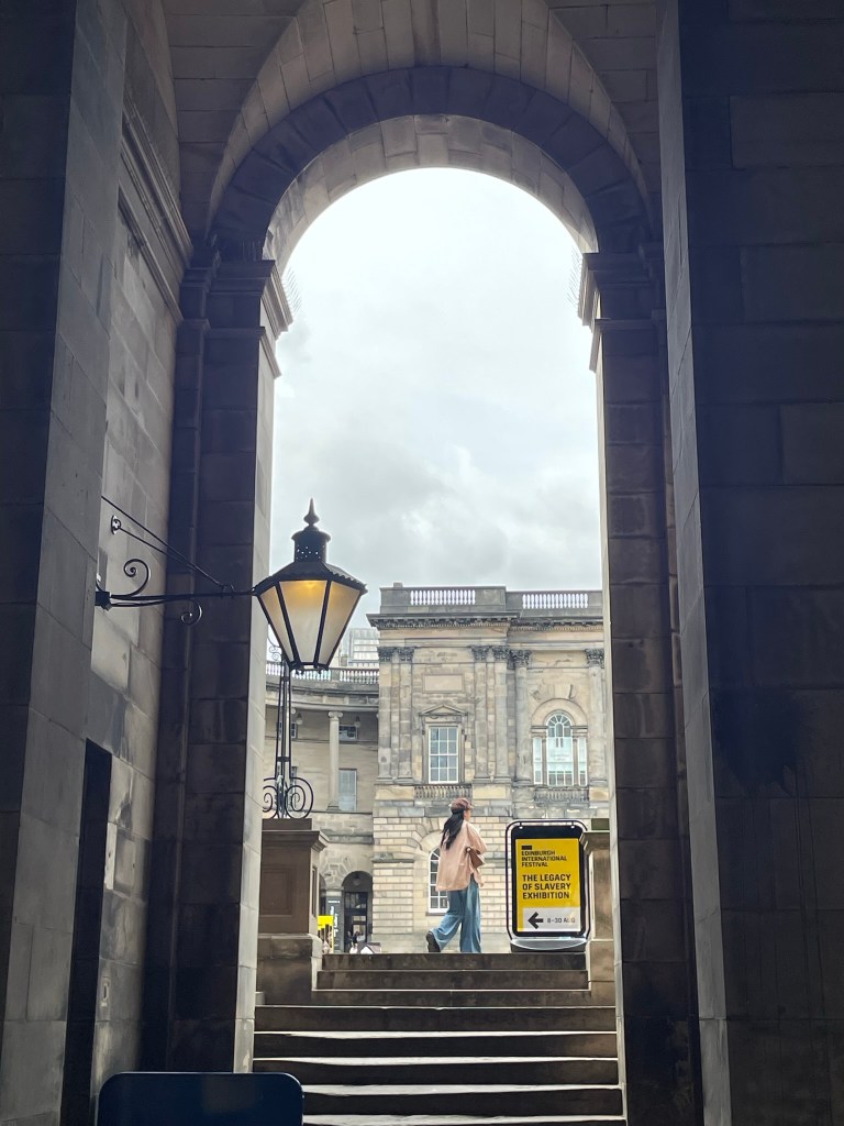 View through an archway with a person walking down steps, lantern hanging to the side, and a historic building visible in the background.