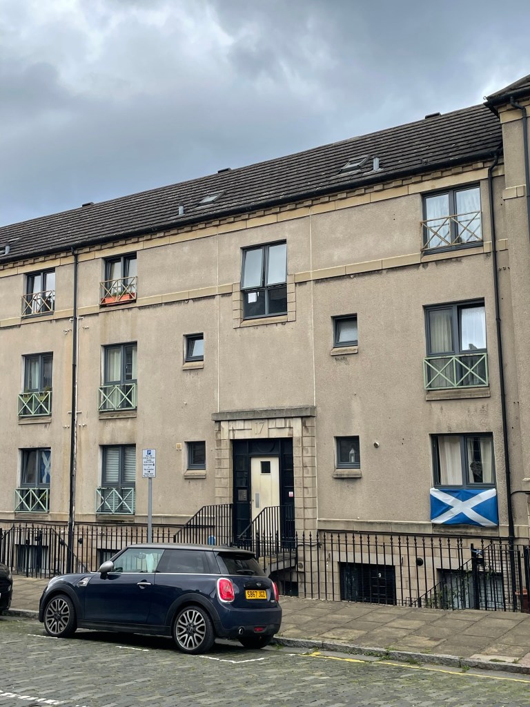 A multi-story residential building with a beige facade, featuring several windows and decorative balconies. A blue car is parked in front, and a Scottish flag is displayed in one of the windows. The sky above is cloudy.