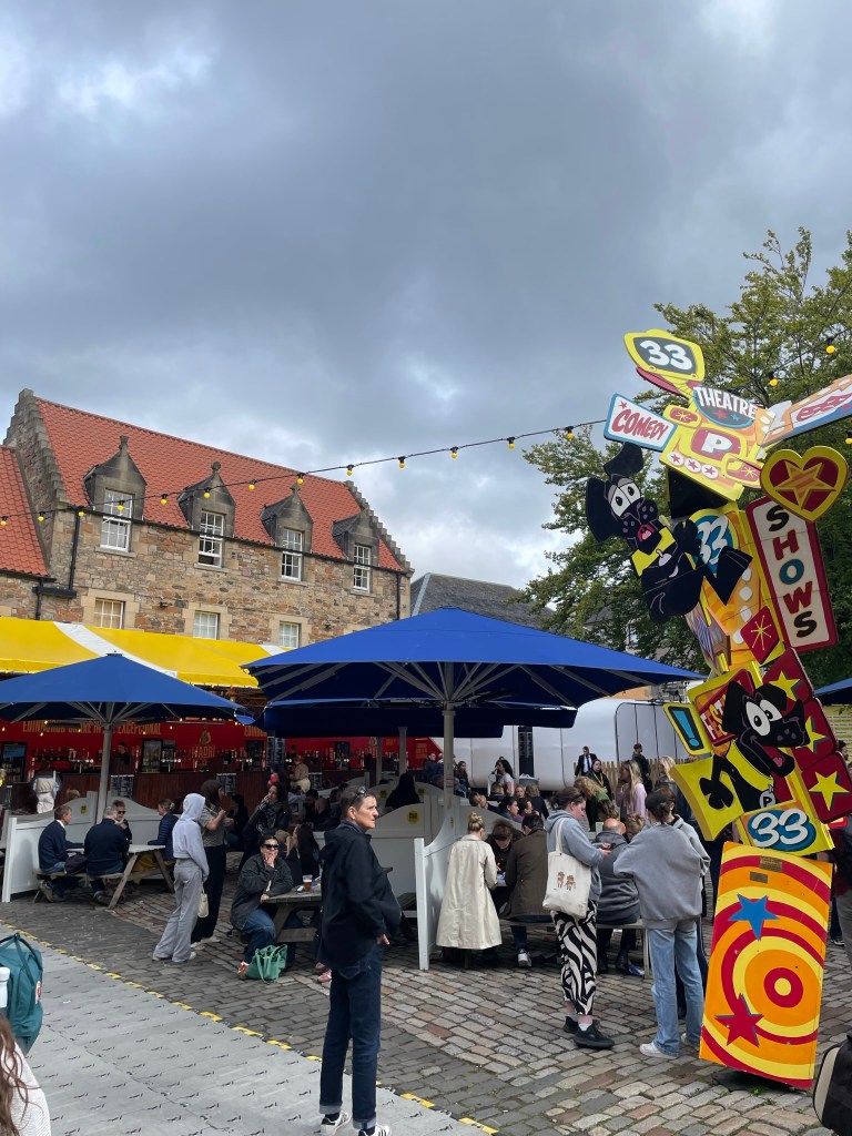 A vibrant outdoor festival scene featuring people seated under large blue umbrellas amid colorful stalls. The backdrop includes an old stone building and a cloudy sky.